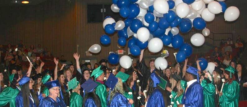 A graduating class at the graduation ceremony. Everyone is is cap and gown and white and blue balloons are falling from the ceiling.