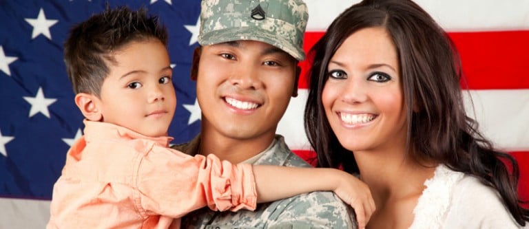 Close up of smiling military family of three. The father is holding their son. They are standing in front of an American flag.